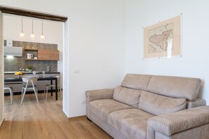 Une cuisine moderne avec des meubles en bois, un coin repas avec une table et des chaises blanches, et un canapé beige dans l’espace salon.