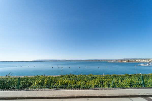 A waterfront view with a clear sky, a paved walkway, and a green fence.