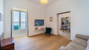 Sala de estar com piso de madeira, uma poltrona azul-esverdeada, um sofá, uma TV fixada na parede e uma cozinha visível por uma porta aberta.