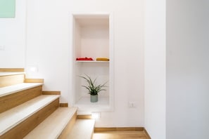 Un escalier menant à un espace intérieur blanc, où l’on aperçoit une étagère sur laquelle sont posés une plante et des vêtements pliés.