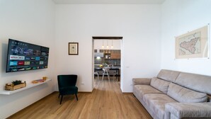 Uma sala de estar moderna com uma TV de tela plana, um sofá, uma poltrona e uma cozinha visível através de uma porta aberta.