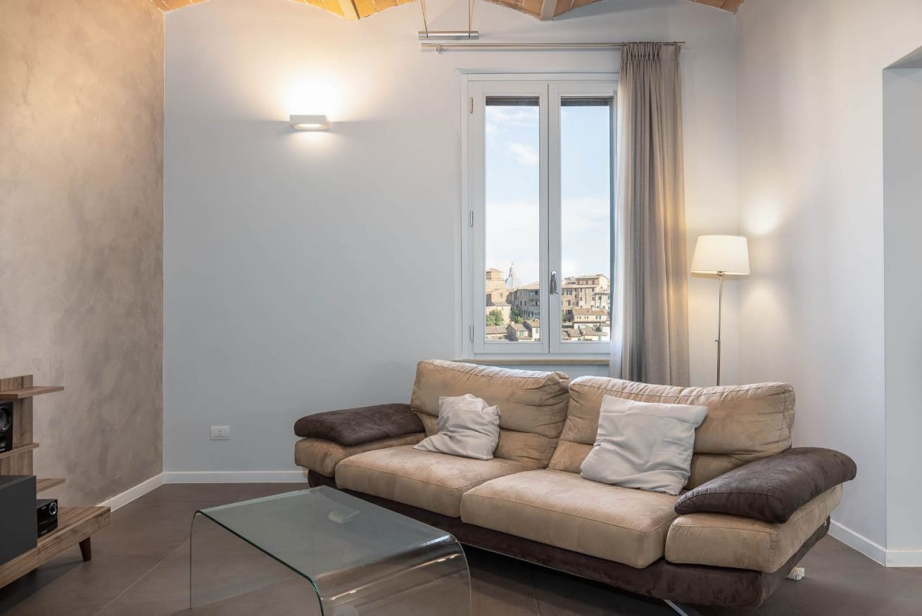 Una sala de estar con sofá, una mesa de centro de vidrio, una lámpara y una ventana con cortinas.