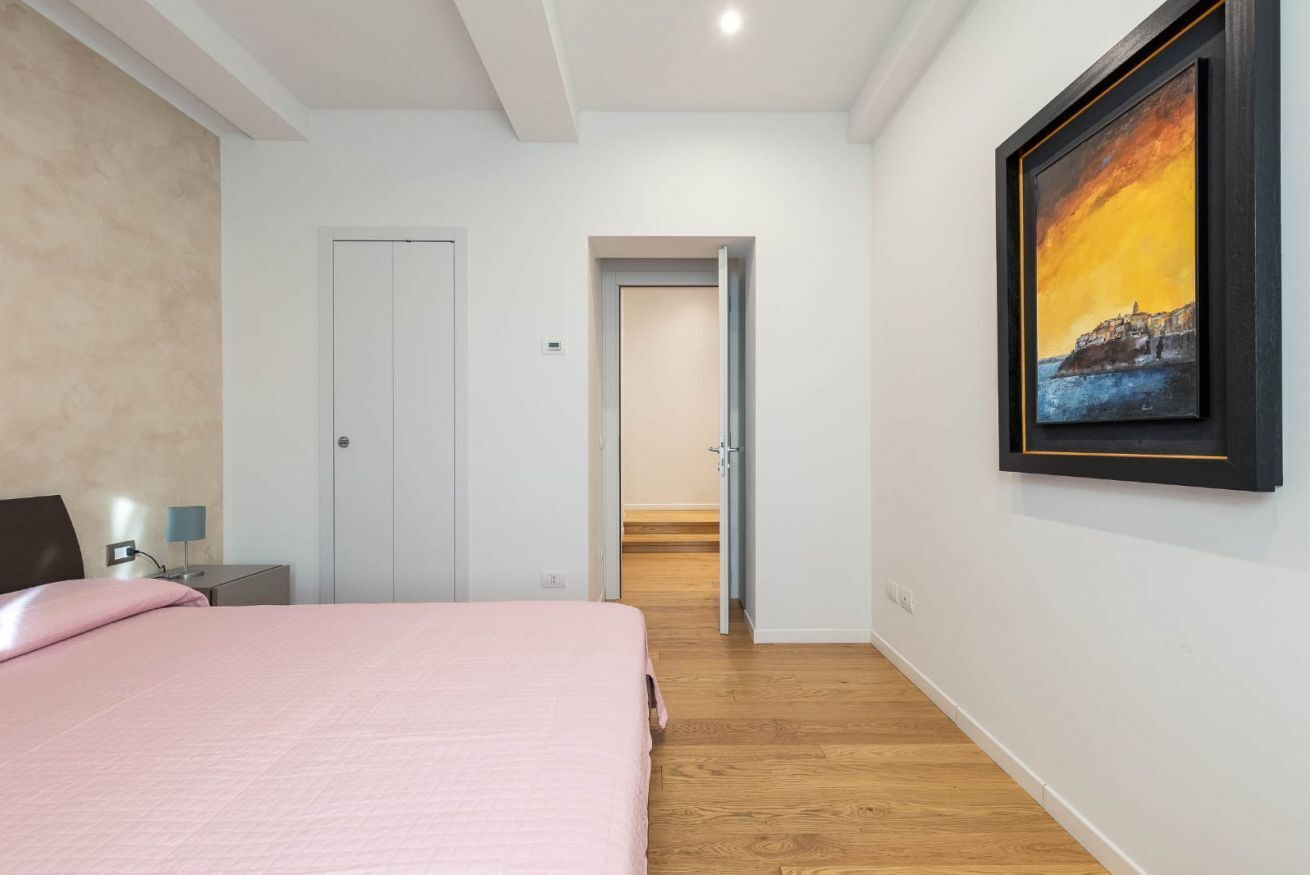A bedroom with a bed, a nightstand, a lamp, a door, and a hallway leading to another room.
