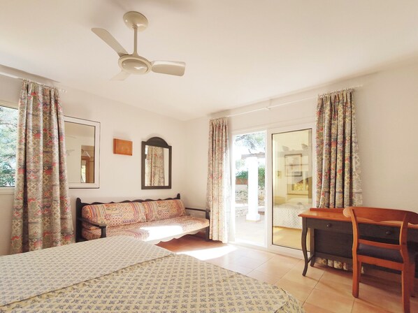 A bedroom with a bed, a desk, a chair, a ceiling fan, a window with curtains, and a sliding glass door leading to an outdoor area.