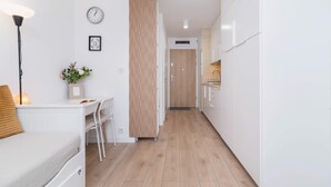 Moderni, hyvin valaistu käytävä, jossa on valkoinen sohva, työpöytä ja ruokailutila. Seinää koristavat kello ja kehystetty taulu.