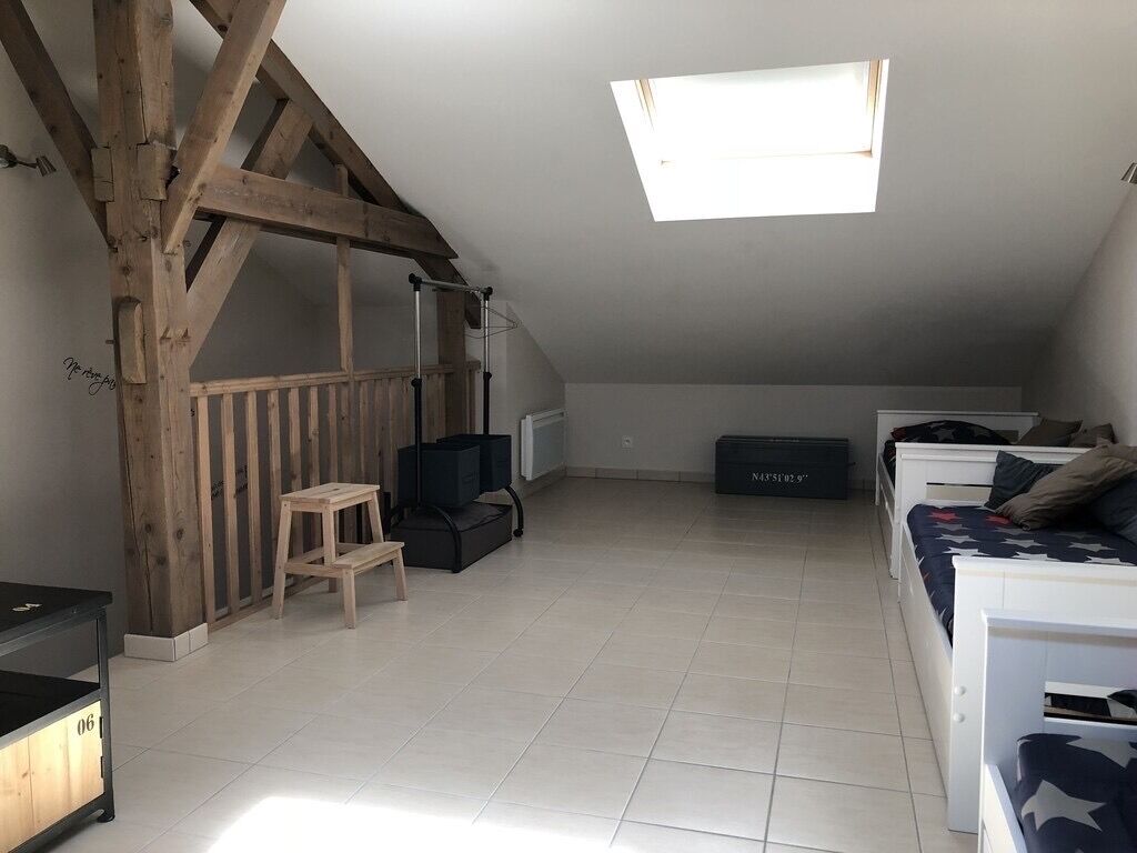 Une pièce avec un plafond en pente, une lucarne, une structure en poutres apparentes, une petite table, un haut-parleur noir, un lit blanc et une boîte de rangement.
