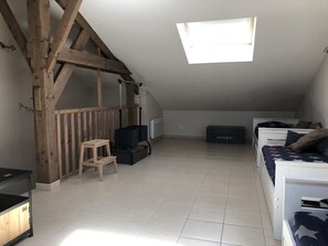 Una habitación con techo inclinado, tragaluz, estructura de vigas de madera, una pequeña mesa, un altavoz negro, una cama blanca y una caja de almacenamiento.