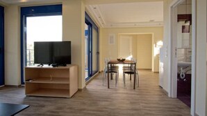 En rummelig opholdsstue med et fladskærms-tv på et træstel, et spisebord med stole, og et stort vindue der giver naturligt lys.