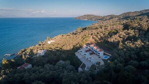 Oversigtsbillede af en kystejendom med flere bygninger, heriblandt en stor villa med swimmingpool, omgivet af frodig vegetation og med udsigt over havet.