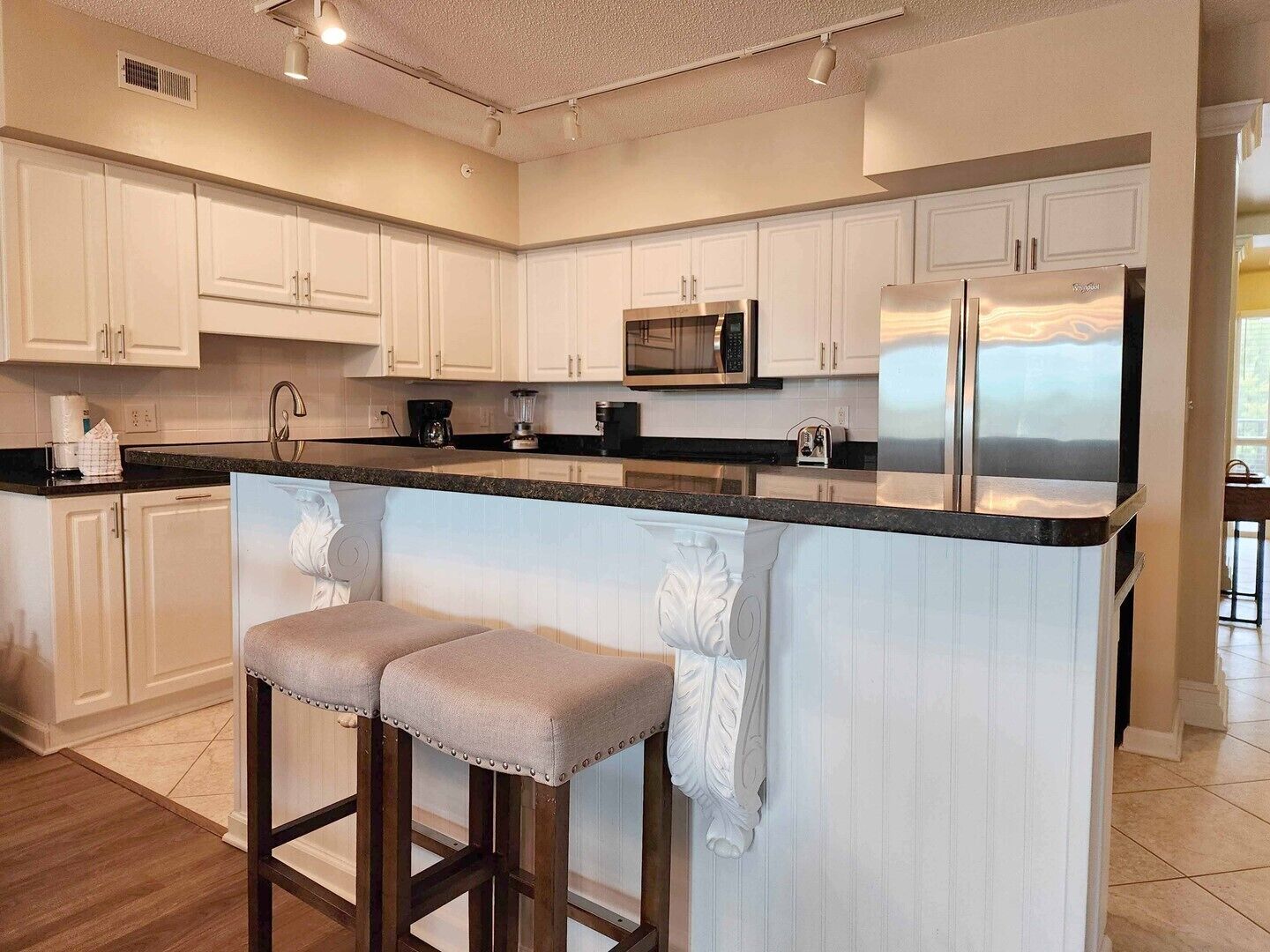 A modern kitchen with white cabinetry, a stainless steel refrigerator, a microwave, and a granite countertop. There are two bar stools with cushioned seats.