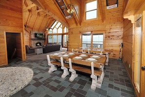 L’intérieur d’une cabane en rondins, avec une grande table à manger, une cheminée en pierre, des poutres en bois et de grandes fenêtres.