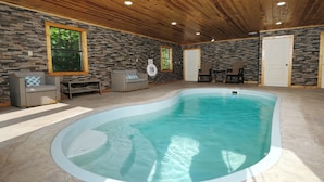Una piscina interior amb parets de pedra i sostre de fusta, envoltada per mobles de vimet i una porta.