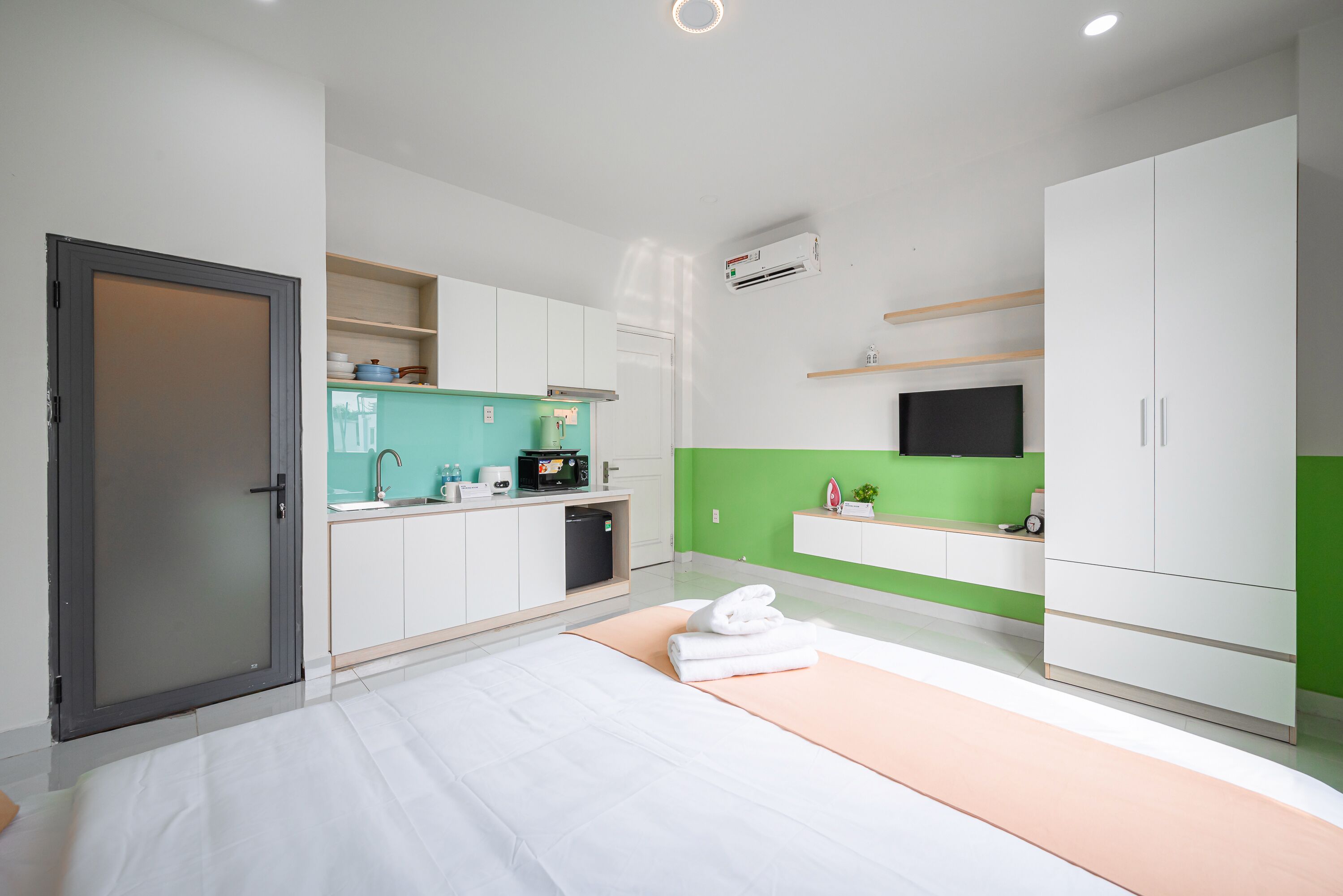 A modern hotel room with a bed, a small kitchenette, a green accent wall, and a large wardrobe.