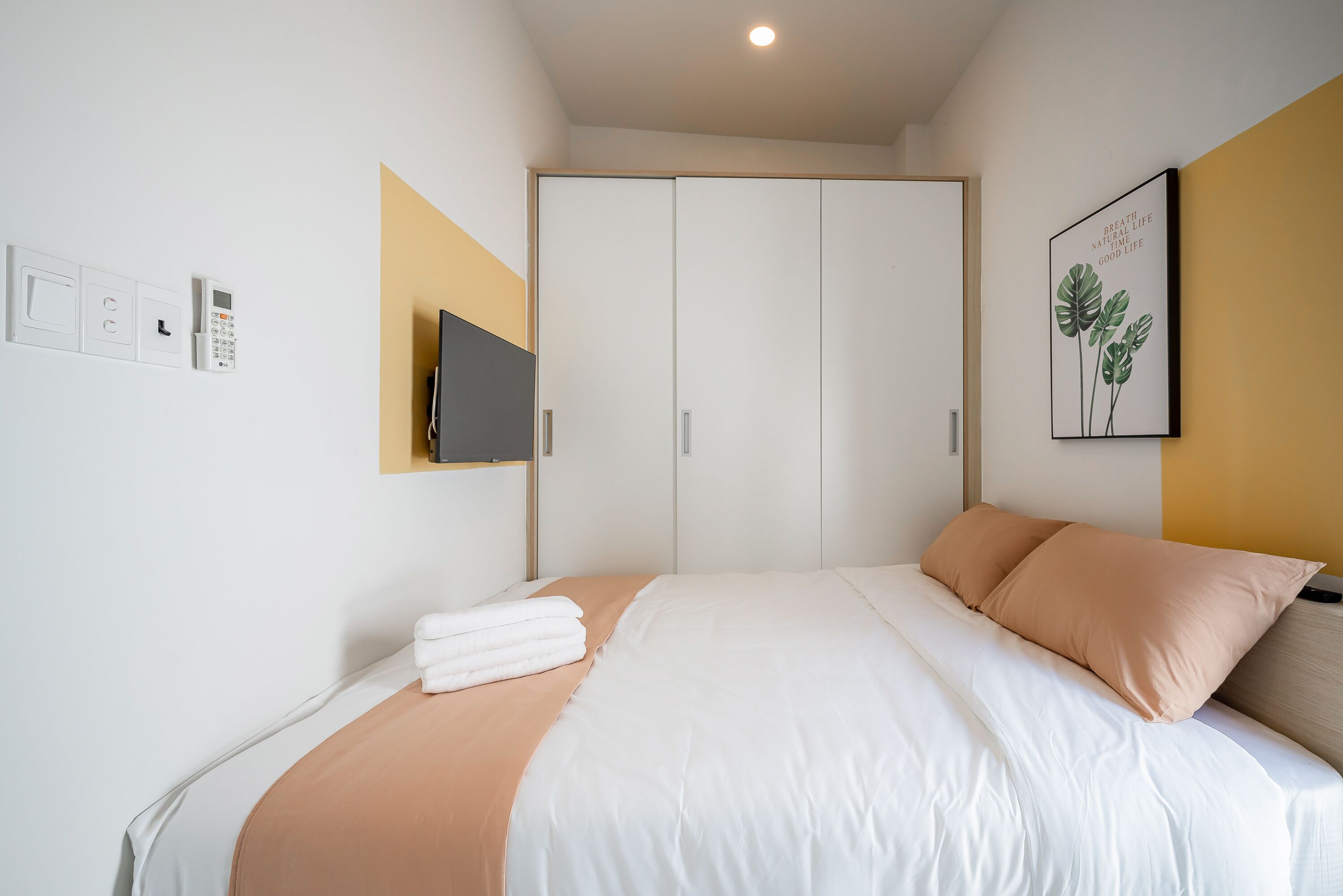 A neatly made bed with white and beige bedding, a flat-screen TV mounted on the wall, and a door with a sliding mechanism.