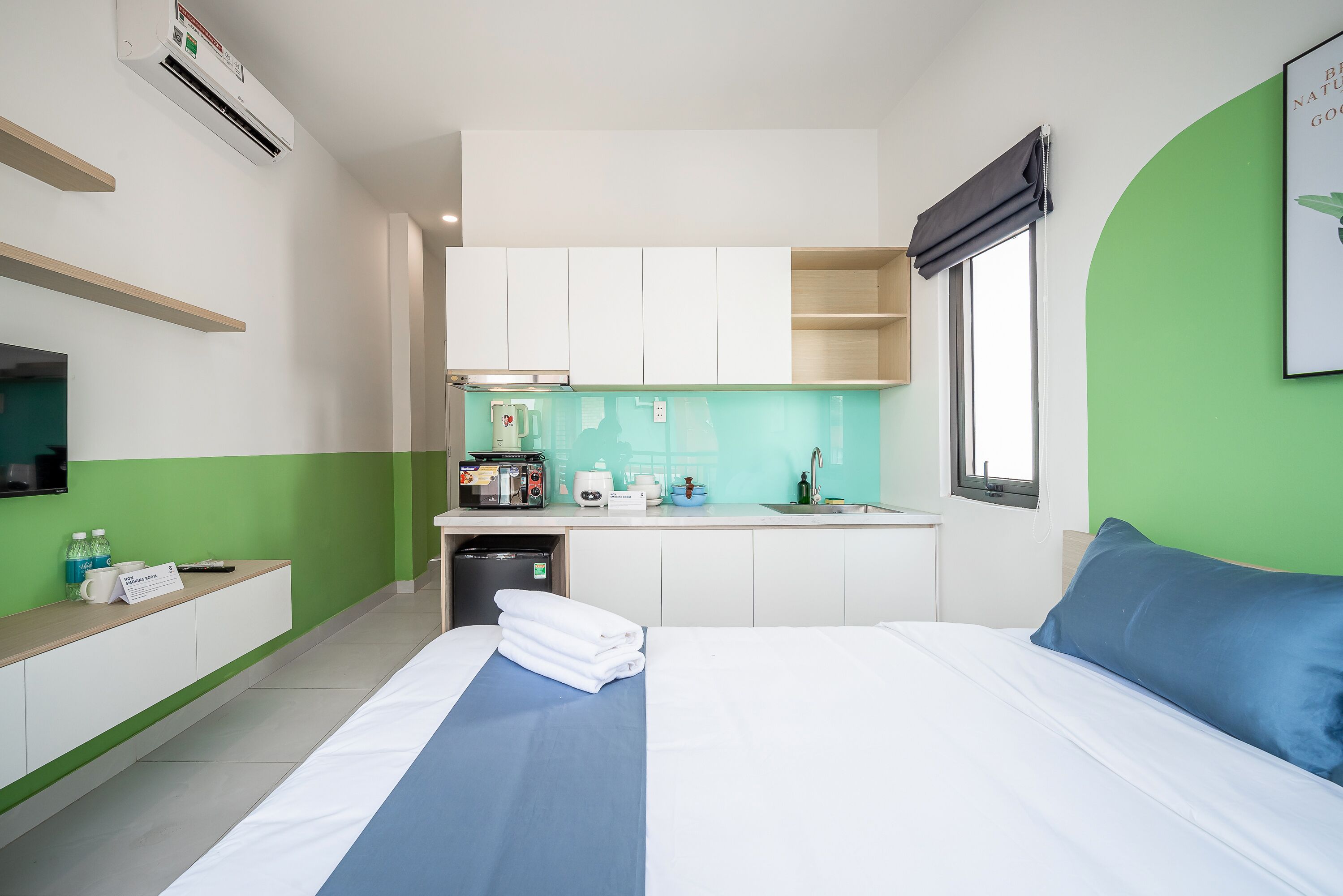 A hotel room with a bed, a small kitchenette, a window, and a green accent wall.