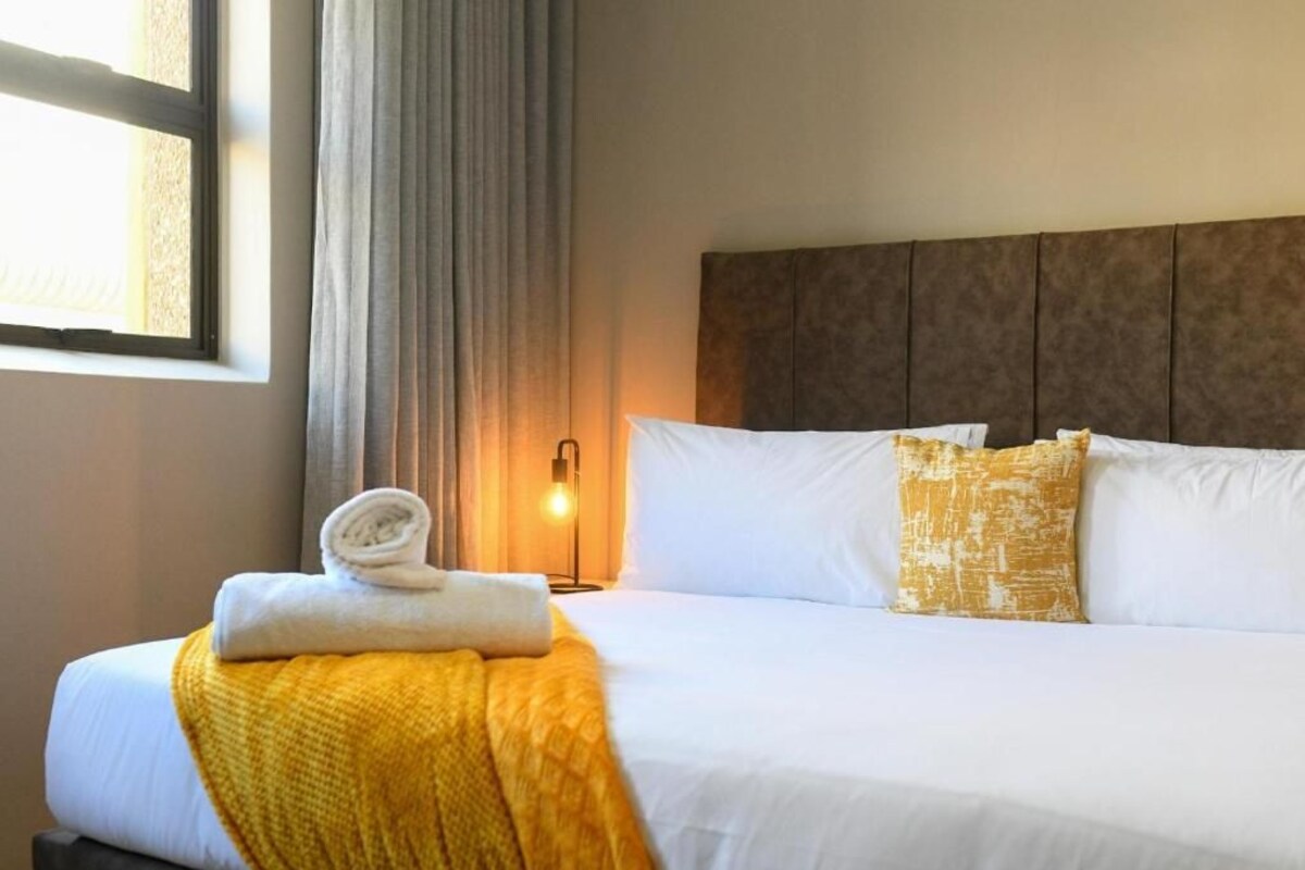 Un lit bien fait, avec du linge de lit blanc, un plaid jaune et un coussin à motifs dorés.