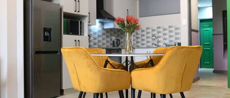 A modern kitchen with a stainless steel refrigerator, a microwave, and a tiled backsplash. There is a dining table with four yellow chairs and a vase with flowers.