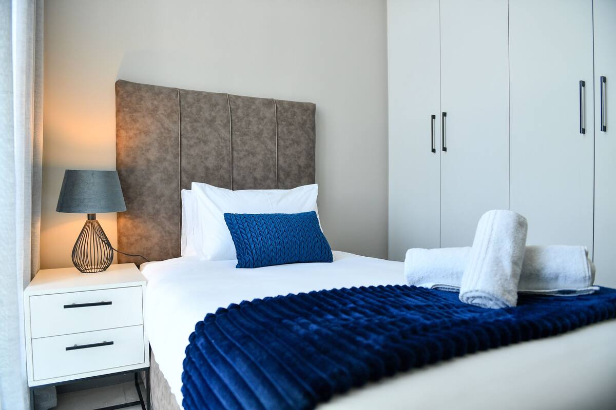 Una cama bien hecha con una manta azul, un almohadón blanco y un almohadón azul decorativo. Se observa una mesita de noche blanca con una lámpara y una toalla doblada. La habitación presenta una paleta de colores neutros, con un cabecero gris y un armario blanco.