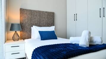 A neatly made bed with a blue blanket, a white pillow, and a blue accent pillow. There is a white bedside table with a lamp and a folded towel. The room has a neutral color scheme with a grey headboard and white wardrobe.