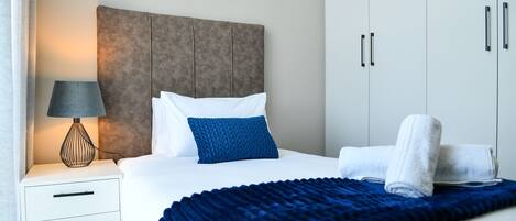 A neatly made bed with a blue blanket, a white pillow, and a blue accent pillow. There is a white bedside table with a lamp and a folded towel. The room has a neutral color scheme with a grey headboard and white wardrobe.