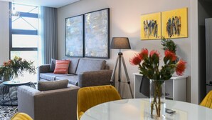 A modern living room with a sofa, armchairs, a coffee table, and a dining table. There are abstract paintings on the wall, a tripod, and a vase with flowers. - Next To Apartment Hotel  (Pretoria)