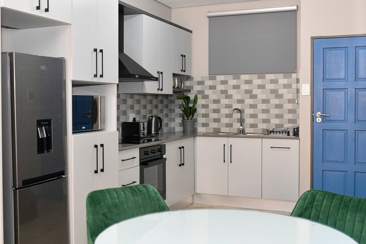 Una cocina moderna con armarios blancos, un refrigerador de acero inoxidable, un microondas y un lavamanos. Se ve una puerta azul y una silla verde.