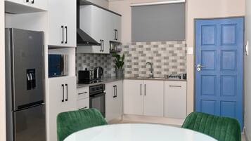 Une cuisine moderne avec des armoires blanches, un réfrigérateur en acier inoxydable, un four à micro-ondes et un évier. On peut voir une porte bleue et une chaise verte.