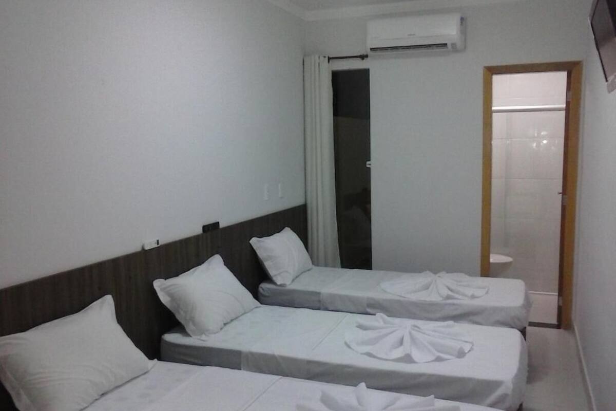 Une chambre d’hôtel avec trois lits simples, chacun recouvert d’une courtepointe blanche et agrémenté d’un coussin décoratif. On y trouve une tête de lit en bois, un mur blanc et un appareil de climatisation fixé au-dessus des lits.