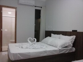 Quarto de hotel com cama, roupa de cama branca, cabeceira de madeira e arranjo de toalhas na cama.