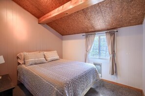 A bedroom with a wooden ceiling, a bed with patterned bedding, a window with curtains, and a radiator.