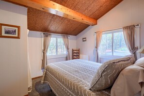 Un dormitorio con techo de madera, cama con ropa de cama estampada, dos ventanas con cortinas y una silla.