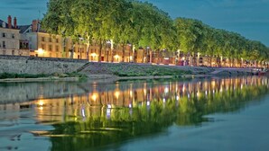 A riverside promenade with buildings along the water, a stone wall, and trees reflecting in the calm water.