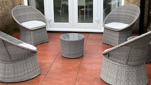 Een terras met rieten meubilair, waaronder twee stoelen en een kleine ronde tafel, voor een glazen deur die uitkomt op een terras.