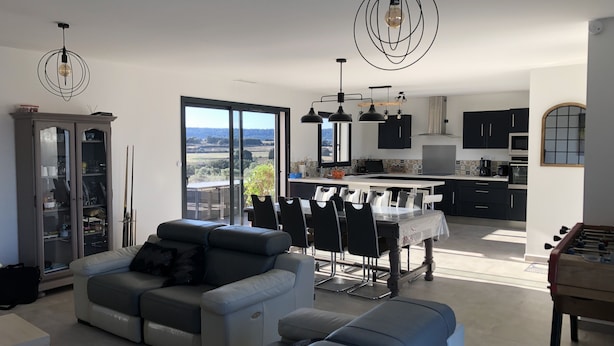 Una sala de estar moderna con sofá, mesa de comedor y sillas. Cuenta con una zona de cocina con armarios y vistas al exterior.