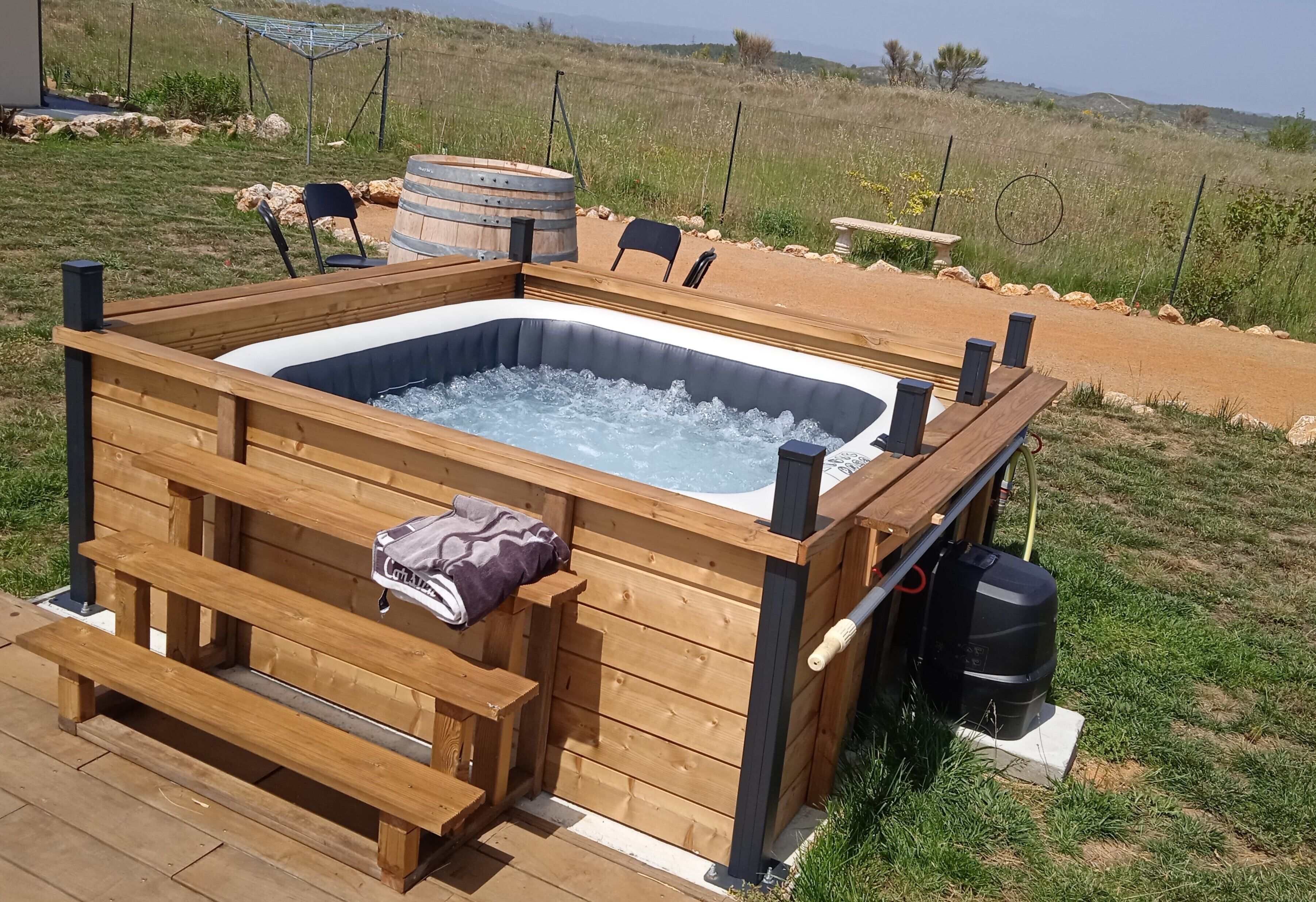 A wooden hot tub with a bench and a table, surrounded by a deck and a grassy area.