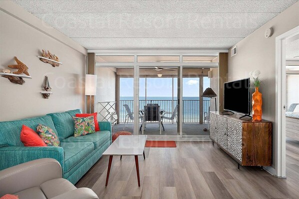 A room with a teal sofa, a coffee table, a TV on a wooden cabinet, and a balcony with a view of the sea.