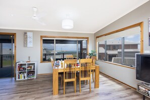 A dining area with a wooden table and chairs, a bookshelf, a television, and large windows offering a view of buildings and a clear sky.