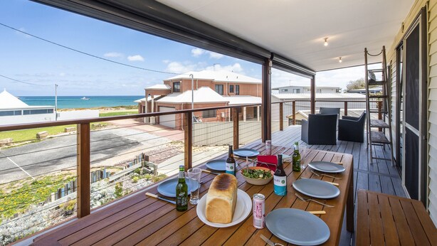Une terrasse en bois avec une table dressée pour le repas, donnant sur une plage et des bâtiments.