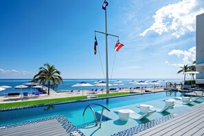 A pool area with a view of the ocean, lounge chairs, umbrellas, and a flagpole with flags.