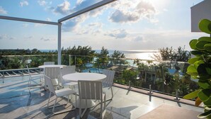 A rooftop terrace with a glass balustrade, white outdoor furniture, and a view of the sea and surrounding landscape. - Rum Point Resort 304 by Grand Cayman Villas (Seven Mile Beach)