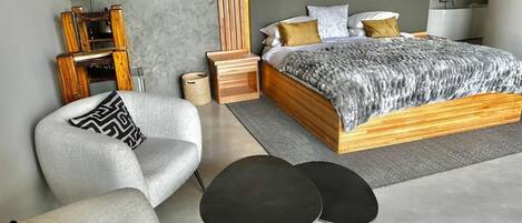 Un dormitorio moderno con estructura de cama de madera, colcha estampada color gris, un sillón gris, una mesita de noche negra y un pequeño tocador de madera.