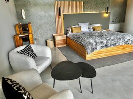 Un dormitorio moderno con estructura de cama de madera, colcha estampada color gris, un sillón gris, una mesita de noche negra y un pequeño tocador de madera.