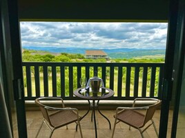 Varanda com duas cadeiras de vime e uma mesa redonda, com vista para uma paisagem verdejante onde se vê uma casa e montanhas ao longe.