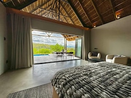 Quarto espaçoso com uma cama grande, área de estar com mesa e cadeiras, e vista para o exterior através de uma porta de vidro.