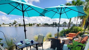 An outdoor patio area with a table and chairs, large umbrellas, and a view of a lake. - Lakeside Cottage. Serene, Tranquil home. 2.mi.(4)mins from the Beach. Sleeps 3.  (Naples)