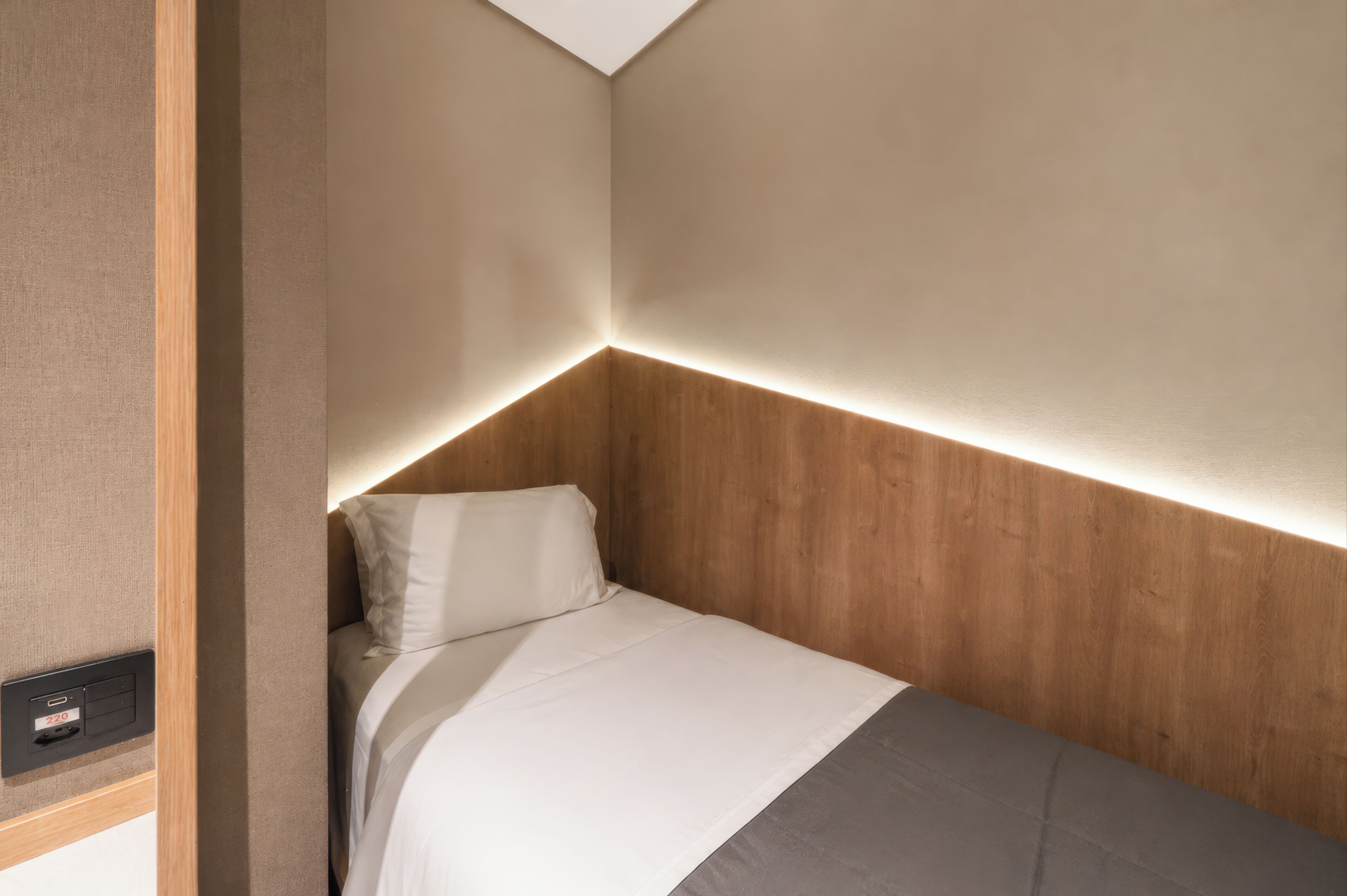 A small, neatly made bed with a wooden headboard and a control panel on the wall.