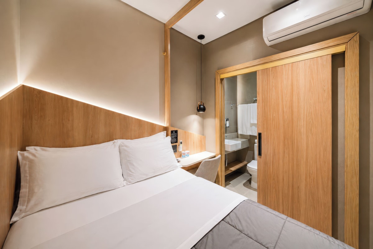 A hotel room with a bed, a desk, a wardrobe, and a bathroom visible through a glass door.