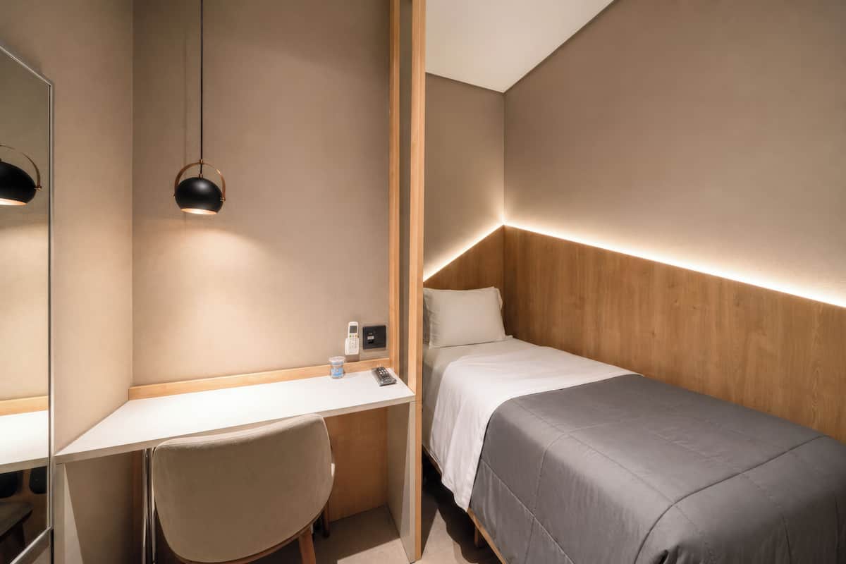 A compact hotel room with a single bed, a desk, a chair, a bedside table, and a mirror.
