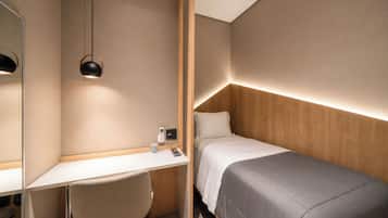 A compact hotel room with a single bed, a desk, a chair, a bedside table, and a mirror.