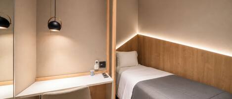 Une chambre d’hôtel de taille modeste, comprenant un lit simple, un bureau, une chaise, une table de chevet et un miroir.